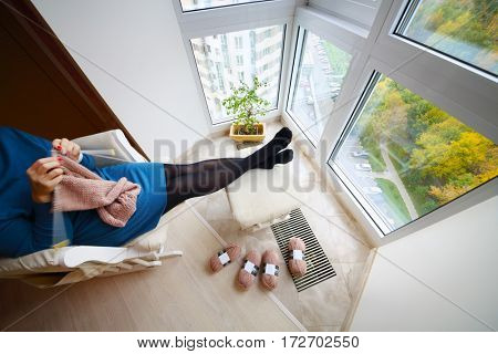Middle-aged knitting woman with feet on banquette sits in armchair near window