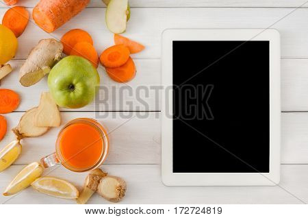 Order online detox cleanse drink, vegetable smoothie ingredients. Natural, healthy juice in glass jar. Carrot, apple, ginger and lemon mix on white wood with tablet screen for copy space, top view