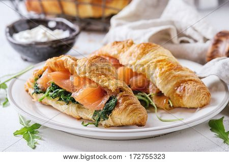 Croissant with smoked salted salmon, spinach and arugula served on white plate with bowl of cream cheese and coffee maker over white and gray concrete background. Close up