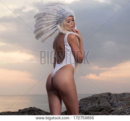 Back view of blond sensual woman wearing white swimsuit and indian feather hatstanding between rocks over beautiful sunset sky and sea background