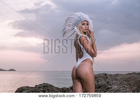 Back view of blond sensual woman wearing white swimsuit and indian feather hatstanding between rocks over beautiful sunset sky and sea background