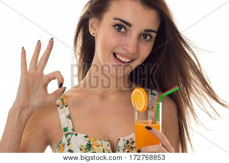 young happy brunette girl in sarafan with floral pattern drinks orange cocktail smiling on camera and showing OK isolated on white