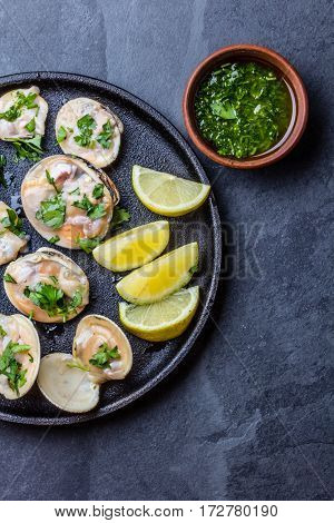 Fresh raw clams with lemon, herbs and white wine on blackiron plate on stone slate background. Top view