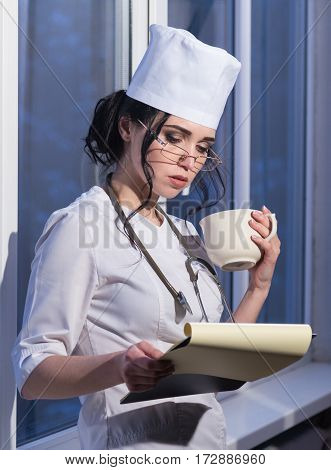 Young and sexy brunette nurse girl in uniform with a stethoscope around neck and a cup of coffee in hands read patient notes