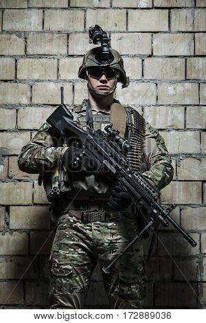 US Army Ranger with machinegun and night vision goggles standing near the wall. Front view portrait
