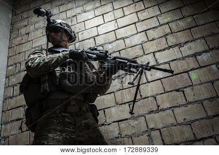 US Army Ranger with machinegun and night vision goggles moving along the wall during mission. Low angle view