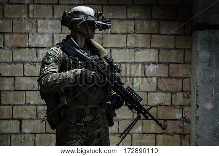 US Army Ranger with machinegun and night vision goggles standing near the wall. Side view profile