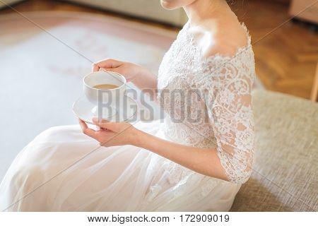 the elegant bride in a white wedding lacy dress holds a tea cup in hand. relaxation before a wedding ceremony. morning tea drinking.