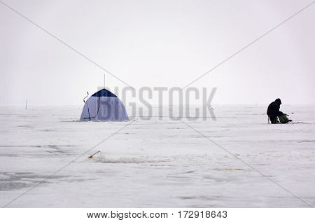 black fisherman in warm clothes sitting on the ice hole tentfishing rod screws winter day Bay sea snow white
