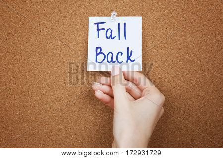 Female hand holding note with phrase FALL BACK on cork board background