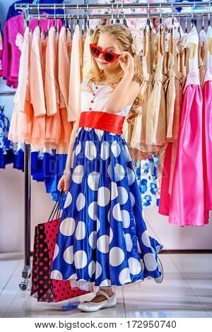 Little fashion-monger girl wearing beautiful pin-up dress and sunglasses goes shopping. Children's fashion. Sale and shopping concept. 