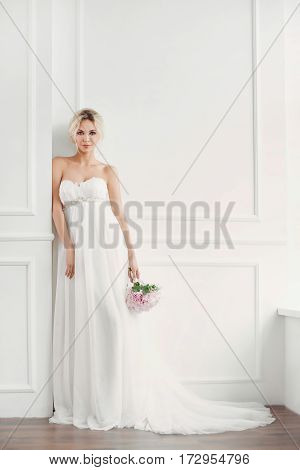 Classical young gourgeous bride. Studio interior fashion shot of fashion smiling model in wedding dress with bouquet of flowers posing in white room. Blonde woman portait with profeshional make-up and hairstyle.