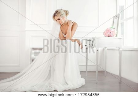 Classical young gourgeous bride. Studio interior fashion shot of fashion model in wedding dress with bouquet of flowers sitting in white room with chair bedside table and mirror. Blonde woman portait with profeshional make-up and hairstyle.