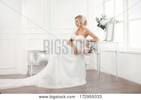 Classical young gourgeous bride. Studio interior fashion shot of fashion model in wedding dress with bouquet of flowers sitting in white room with chair bedside table and mirror. Blonde woman portait with profeshional make-up and hairstyle.