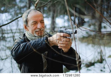 An elderly man with a gun in the woods in early spring. Man dressed in a black jacket and scarf. He holds the gun in both hands and aiming into the distance