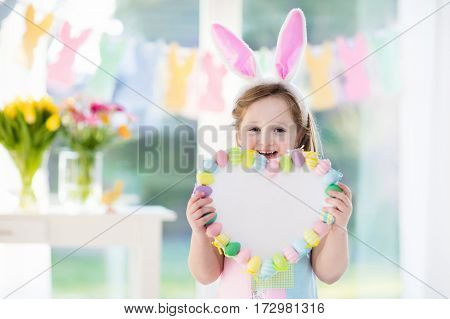 Happy little girl in bunny ears holding a heart board with colorful Easter eggs. Kids celebrate Easter. Children having fun on Easter egg hunt. Pastel bunny banner and flowers. Space for your text.