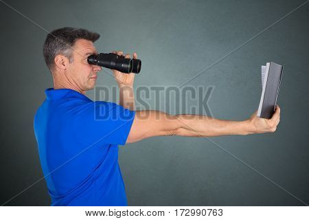 Matured Man With Hyperopia Eye Disorder Reading A Book Using Binocular On Gray Background