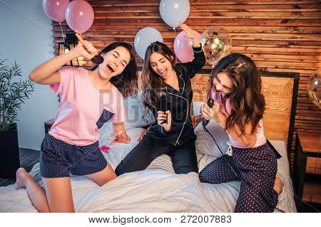 Playful And Nice Young Women Stnding On Knees On Bed In Room. They Pretend Singing In Microphones. M