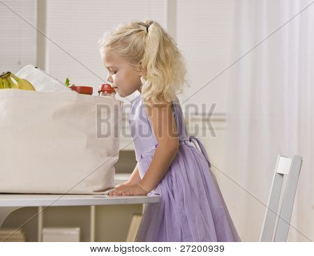 Молодая девушка peeking в мешок бакалеи на кухонном столе.  Она смотрит от t