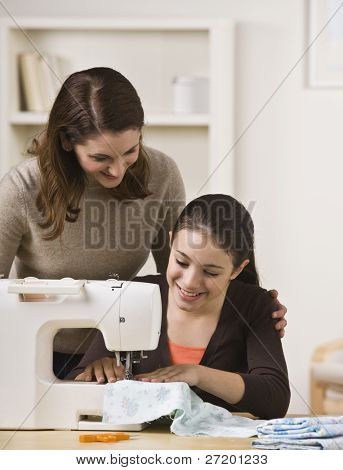 A beautiful teenage girl sewing on a sewing machine while her mother watches over her shoulder.  They are smiling.  Vertically framed shot.