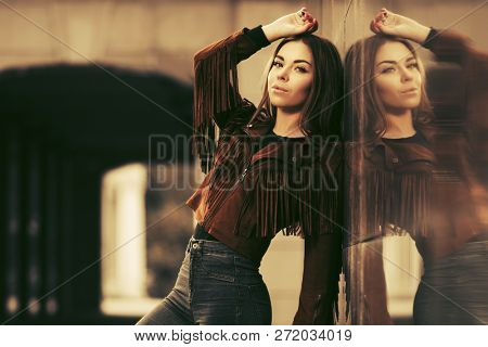 Happy young woman standing at the wall on city street  Stylish fashion model wearing brown leather fringe suede jacket and dark blue jeans