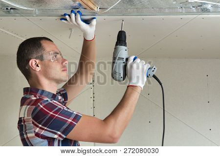 Young Man In Goggles Fixing Drywall Suspended Ceiling To Metal Frame Using Electrical Screwdriver On