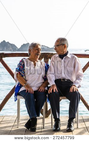 Elderly Couple Looking At Each Other Laughing. Elderly Couple With The Sea Background.
