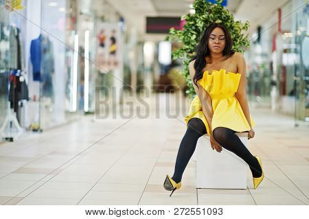 Stylish African American Woman At Yellow Dreess Posed At Mall And Sitting On Bench Inside.