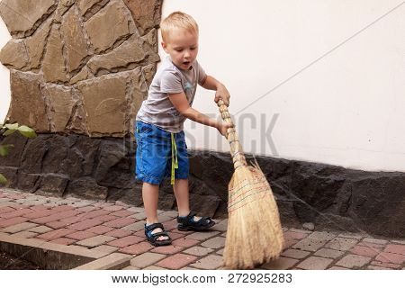 Pretty Caucasian Baby Boy With Blonde Hair Sweeps The Yard With A Broom. Outdoors, Daylight.