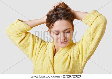 Studio Shot Of Tender Concetrated Woman Focused Down, Gathers Hair, Wears Domestic Robe, Enjoys Week