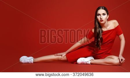 Beautiful brunette girl posing at studio in red dress and white sneakers. Red background. Sport chic style.