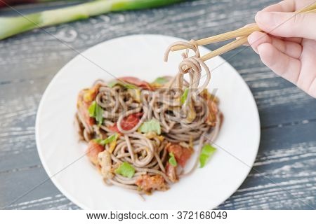 Brown Noodles Or Soba On Chopsticks In Girls Hand. Fresh Soba On The White Plate With Chicken And Ve