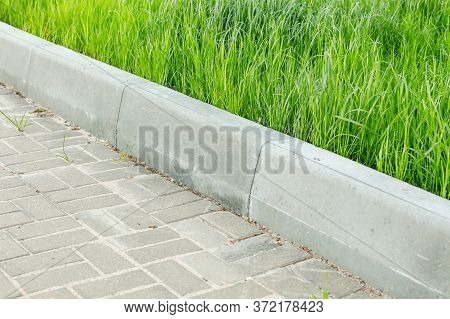 A Curbstone Separates The Pedestrian Zone From The Green Tall Grass Of The Lawn. City Improvement