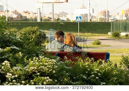 Loving Couple Of Young People, A Girl With Red Hair And A Guy In A Plaid Shirt Are Sitting On A Benc