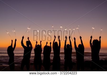 A multi-ethnic group of friends enjoying their time together on a beach during sunset, standing in a row, holding sparklers, raising their arms