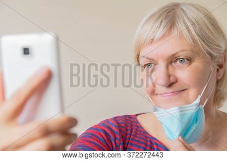 Mature Woman Taking Selfie Holding Medical Mask In Hand. Elderly Woman Taking Off Medical Face Mask 