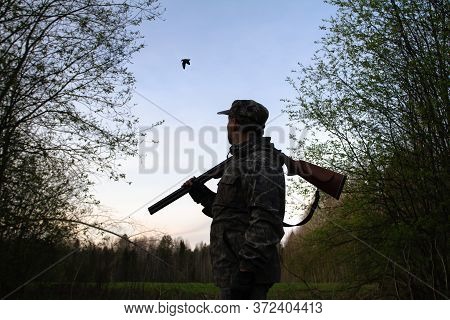 A Hunter With An Unloaded Shotgun On His Shoulder Stands In The Dusk In A Forest Clearing And Sees A