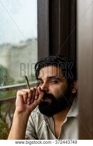 A Bearded Jobless Indian Man Wearing Kurta Pyjama Smoking Weed Rolled In A Paper. He Is Staring At C