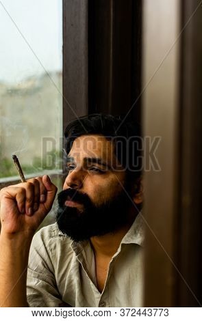 A Bearded Jobless Indian Man Wearing Kurta Pyjama Smoking Weed Rolled In A Paper. He Is Staring At T