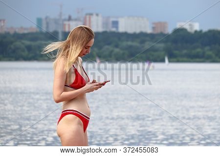 Moscow, Russia - June 2020: Blonde Girl In Red Bikini Standing On A Beach With Smartphone In Hands O