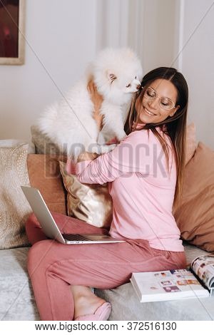 A Girl In Pajamas At Home Is Working On A Laptop With Her Dog Spitzer, The Dog And Its Owner Are Res