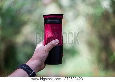 Male Hand Holds A Thermomug. Red Thermomug On A Background Of Greenery