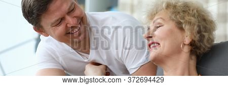 Elderly Woman Reads Book At Home, Next Adult Son. Home Quarantine Upon Contact With Patient. Strict 