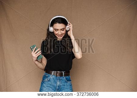 Photo of young brunette woman smiling and listening to music with headphones isolated over beige background