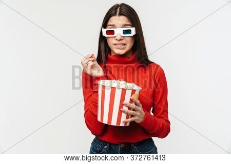 Photo of uptight brunette woman wearing 3D glasses holding popcorn bucket isolated over white background