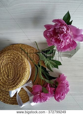 Two Peonies Lay On A Wicker Straw Hat On A Light Wooden Background. One Peony Is Standing In A Trans