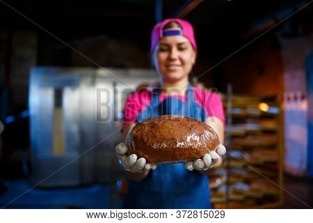 A Baker Girl Takes Hot Bread In A Bakery Against The Background Of Shelving With Bread. Industrial P