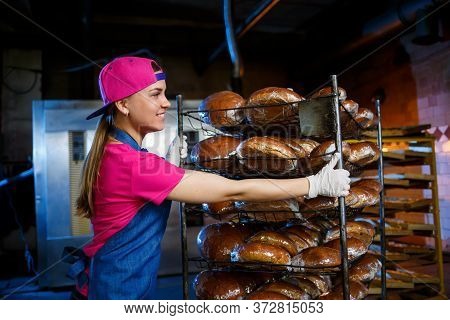 A Baker Girl Takes Hot Bread In A Bakery Against The Background Of Shelving With Bread. Industrial P