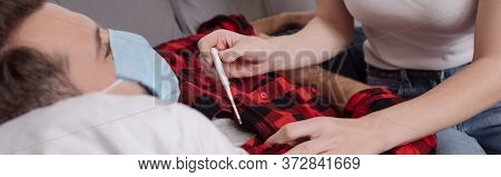 View Of Young Woman Holding Thermometer Near Sick Boyfriend Lying On Sofa In Medical Mask, Panoramic