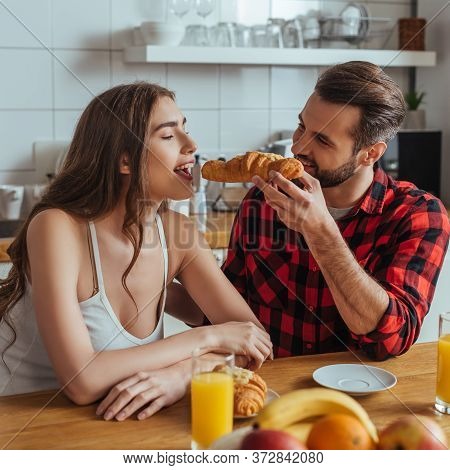 Handsome Man Feeding Attractive Girlfriend With Delicious Croissant Near Orange Juice And Fresh Frui